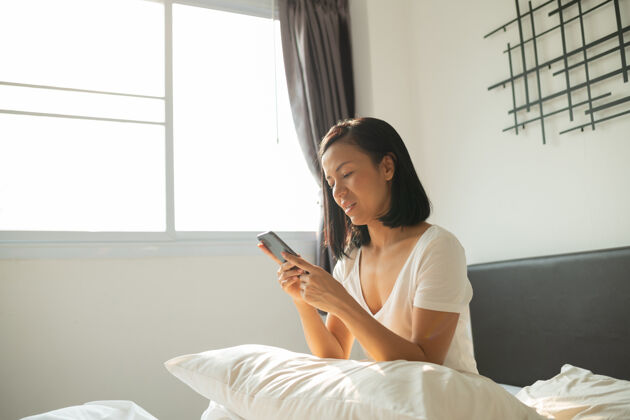 房间微笑迷人的亚洲女性在白色床上使用平板智能手机小睡睡衣躺下