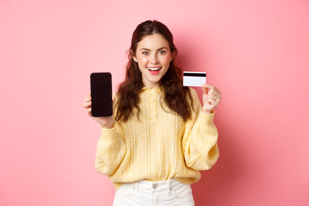候选人科技和网上购物年轻迷人的女模特展示空的智能手机屏幕和塑料信用卡 展示银行账户或应用程序 站在粉红色的墙上时尚电话乐观
