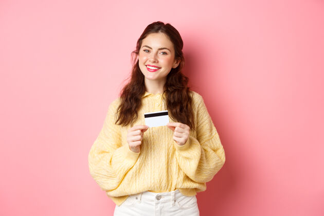 快乐美丽的女模特想着购物 拿着塑料信用卡微笑着 站在粉色的春日墙上复制空间水平女人女孩