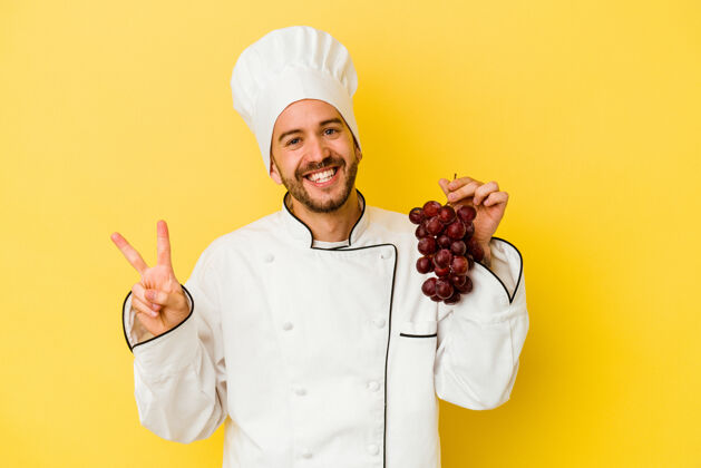 烹饪年轻的白人厨师手拿着黄色背景上孤立的葡萄 快乐而无忧无虑地用手指展示着和平的象征和平欢乐赢家