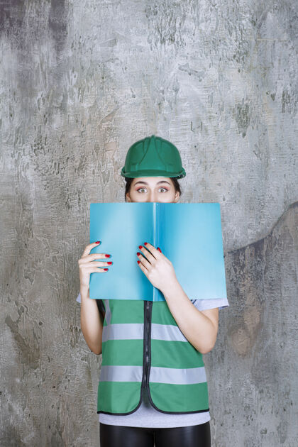 工人身穿绿色制服 头戴安全帽的女工程师 手里拿着一个蓝色的项目文件夹 后面藏着一张脸压力害怕年轻人