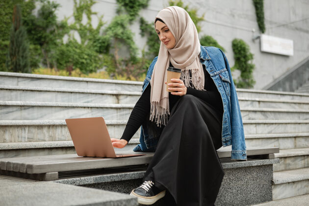 阿拉伯语现代时尚的妇女戴着头巾 穿着牛仔夹克和黑色长袍坐在城市街道上用笔记本电脑工作城市民族自营职业