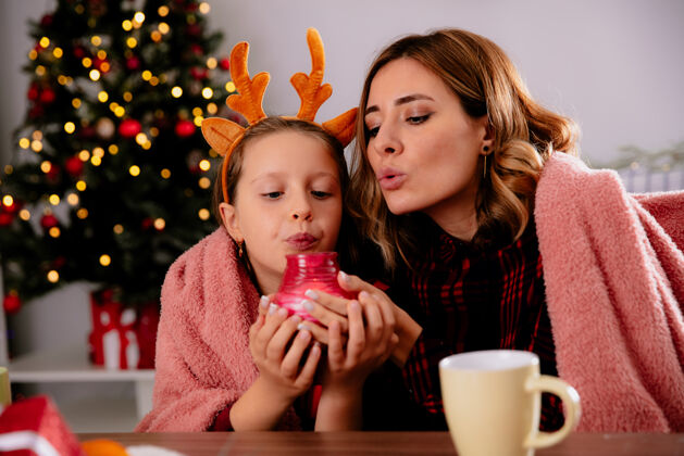 房子高兴的母亲和女儿抱着毯子 吹着蜡烛坐在桌旁 享受着圣诞节的时光在家里抱着坐着吹