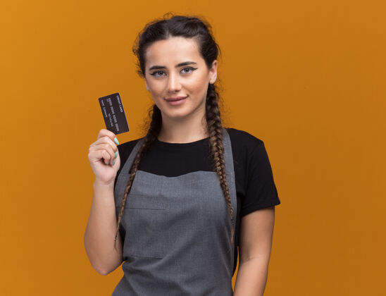 年轻人高兴的年轻女理发师穿着制服拿着信用卡隔离在橙色的墙上女性衣服理发师