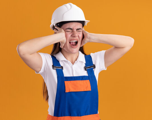脸愤怒地闭上眼睛 年轻的建筑女工穿着制服 把手放在耳朵上 隔离在橙色的墙上感觉封闭年轻