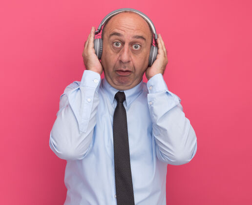 姿势惊讶的中年男子穿着白色t恤 打着领带 把耳机孤立在粉红色的墙上人人耳机