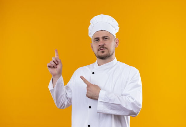 年轻自信的年轻帅哥厨师身着厨师制服 孤立地指着橙色的墙上帅气厨师厨艺
