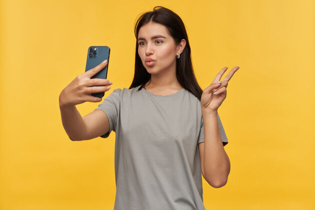 黑发迷人的顽皮的黑发年轻女子在灰色T恤显示和平标志发送一个吻 使鸭脸和自拍使用智能手机越过黄墙模特年轻人站立