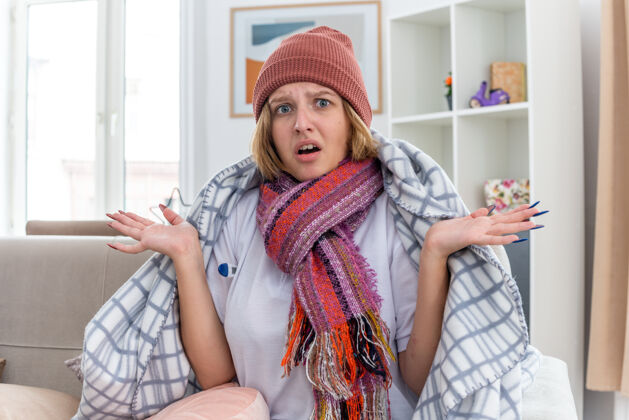 帽子不健康的年轻女子 戴着暖和的帽子 裹着毯子 看起来不舒服 生病 感冒和流感 体温计发烧 看起来很困惑 坐在客厅里的沙发上生活沙发痛苦