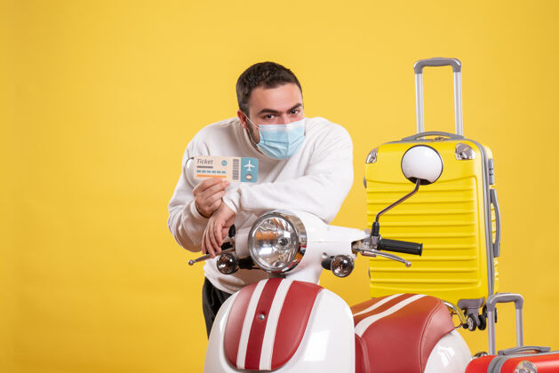 运动员旅行概念的俯视图 戴着医用面罩的人站在摩托车旁边 黄色手提箱在上面 手里拿着车票票运动人