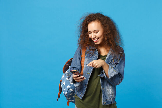 短信年轻的非洲裔美国女孩十几岁的学生在牛仔服装 背包举行电话隔离在蓝色的墙壁上手机电话卷发