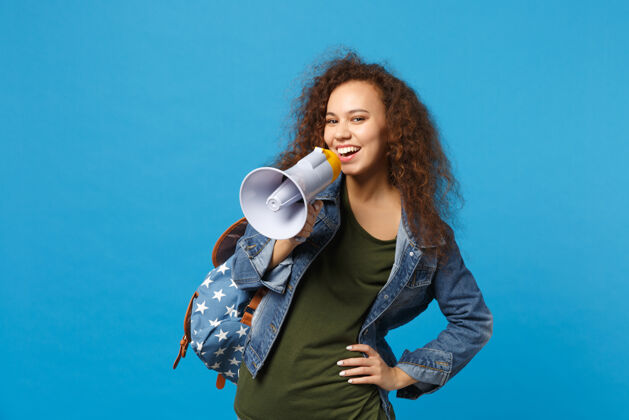 扬声器年轻的非洲裔美国少女 穿着牛仔服 背着背包 把扩音器孤立地放在蓝色的墙上扩音器广播音频