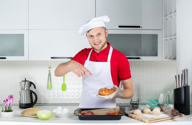 男厨师白色厨房里 年轻的面带微笑的男厨师手持并指着一个新鲜出炉的糕点 戴着支架成年人持有人烹饪