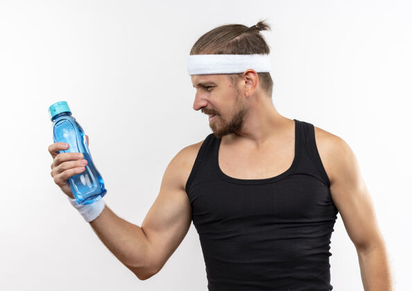 运动自信的年轻帅气的运动型男人戴着头带和腕带拿着水瓶看着隔离在白墙上瓶子自信抱着