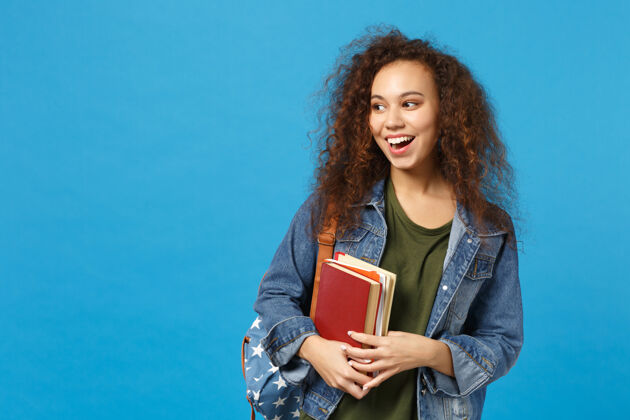 女穿着牛仔服 背着书包的年轻女学生把书孤立地放在蓝色的墙上非洲快乐学者