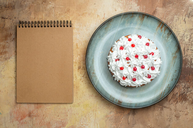 笔记本上图：蓝色盘子上装饰着奶油和葡萄干的美味蛋糕 彩色桌子上的螺旋笔记本盘子桌子古董