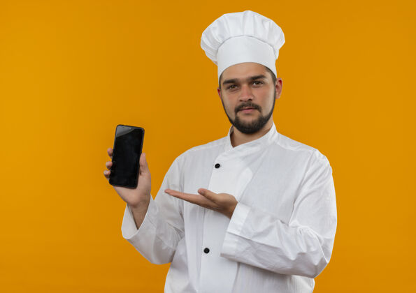 男性自信的年轻男厨师身着厨师制服 用手指着隔离在橙色墙上的手机信心厨师手机