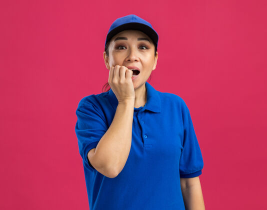压力穿着蓝色制服 戴着帽子的年轻女送货员紧张地站在粉红色的墙上咬指甲帽子年轻制服