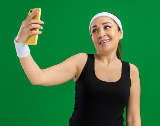 头带戴着头带和臂章的年轻健身女士开心而积极地微笑着用智能手机站在绿色的墙上自拍移动年轻积极