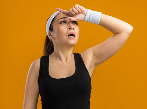 女人戴着头巾和臂章的年轻健身女士抬起头来 手放在头上 站在橘色的墙上 疲惫不堪 劳累过度过度劳累头带头