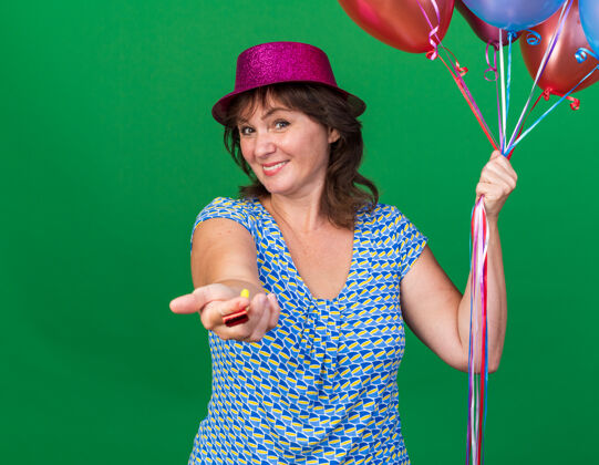 年龄戴着派对帽的中年妇女手持五颜六色的气球 面带微笑地吹着口哨站在绿色的墙上庆祝生日派对中间举行帽子