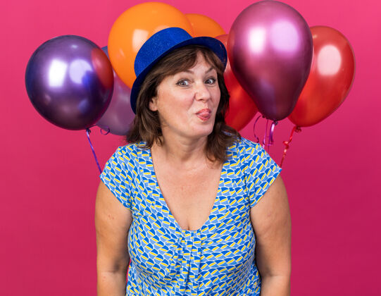 举行滑稽开朗的中年妇女 戴着派对帽 手持五颜六色的气球 伸出舌头 站在粉色的墙上庆祝生日派对生日五颜六色舌头