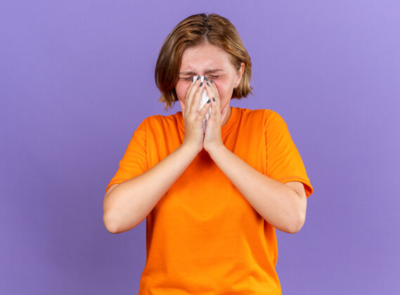 感觉不健康的年轻女子穿着橙色t恤 感觉很糟糕 流鼻涕感冒了 站在紫色的墙上打喷嚏站着抓到吹
