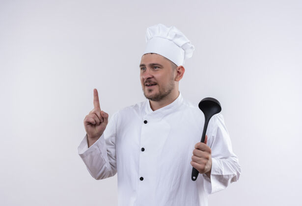 烹饪印象深刻的年轻帅哥厨师在厨师制服拿着勺子 抬起手指看着隔离在白墙上的一面手指提高印象