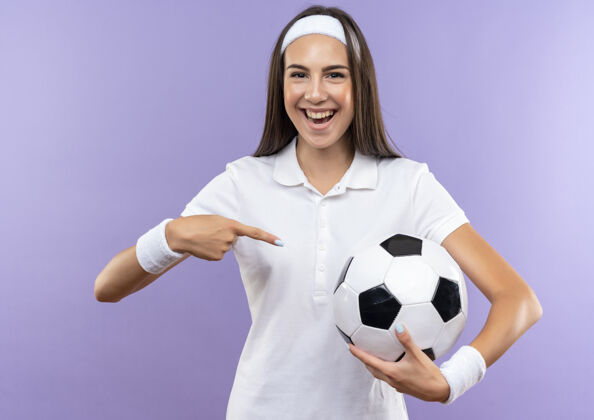 穿快乐漂亮的运动女孩戴着头带和腕带拿着和指着足球隔离在紫色的墙壁头带运动球