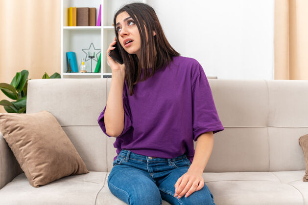 电话一个穿着休闲服的年轻女孩坐在客厅里的沙发上 一边用手机聊天 一边显得很烦躁房间生活年轻