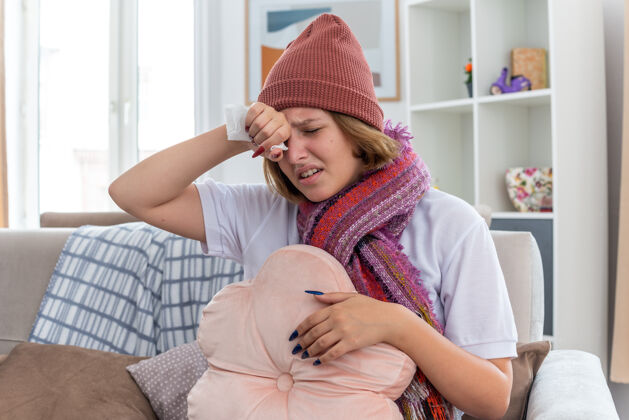 生活不健康的年轻女子戴着暖和的帽子 戴着围巾 看起来不舒服 摸着额头 发烧 头痛 患感冒和流感 坐在明亮的客厅沙发上女人坐着帽子