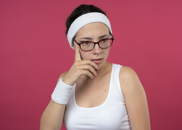 手体贴的年轻运动女孩戴着眼镜 戴着头带和腕带 手放在下巴上 往下看运动羽绒穿