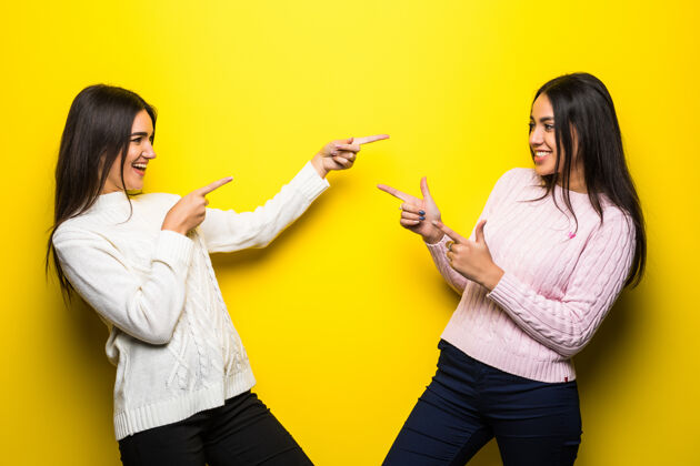 享受两个穿着毛衣的快乐女孩的肖像 在黄色的墙上互相手指女人手势微笑