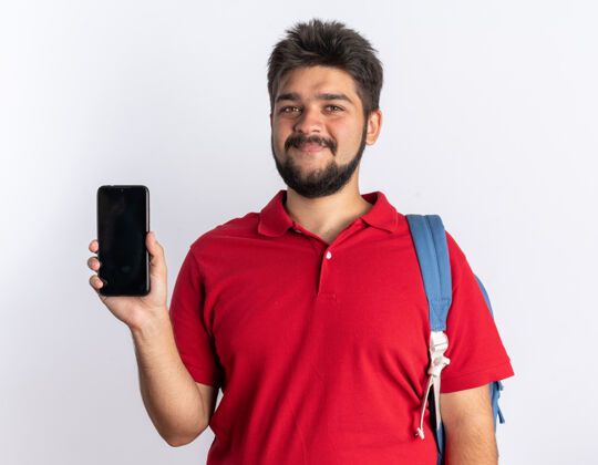 年轻年轻的留着胡子的学生 穿着红色马球衫 背着背包 手里拿着智能手机 站着学生背包马球