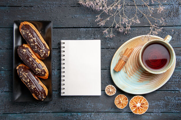 桌子俯视图：矩形板上放着巧克力蛋糕 黑色木桌上放着一本笔记本和一杯茶 柠檬干和肉桂茶杯子顶部