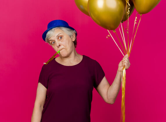 帽子震惊的老妇人戴着聚会帽手持氦气球吹着粉红色的口哨吹粉色口哨