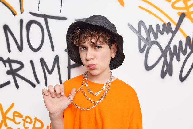 垃圾十几岁的街头艺术家戴着帽子 穿着橙色t恤和项链 对着墙上的涂鸦摆出不满的表情板姿势青年
