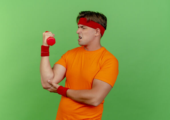 姿势紧张的年轻英俊的运动型男子戴着头带和腕带举起哑铃把手放在肘部隔离在绿色的墙上腕带年轻肘