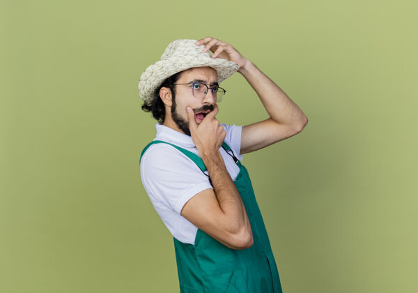 胡须年轻的留着胡须的园丁 穿着连体衣 戴着帽子 站在浅绿色的墙上 一边看着一边惊讶和惊讶花园市民连身衣