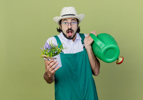 年轻人年轻的留着胡须的园丁 穿着连体衣 戴着帽子 手里拿着水罐和盆栽植物 站在浅绿色的墙上 惊讶地看着前方帽子人惊喜