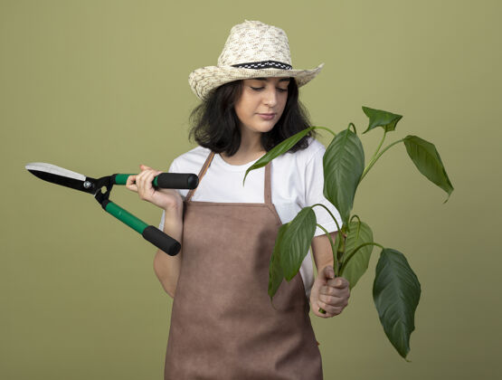 姿势自信的年轻黑发女园丁穿着制服 戴着园艺帽 手持园艺剪 看着橄榄绿墙上孤立的植物帽子花园脸