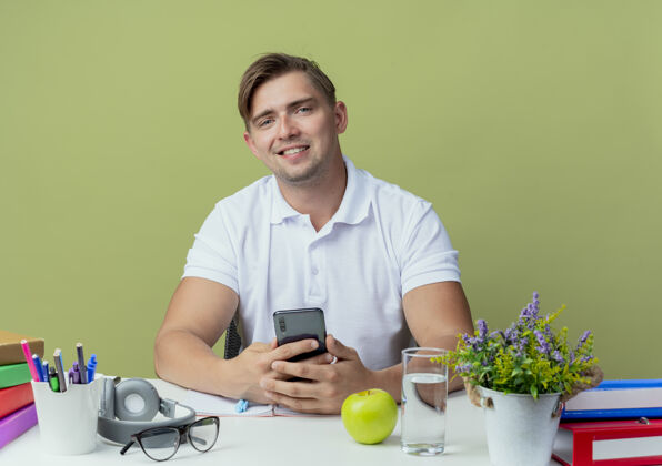 坐着微笑的年轻英俊的男生坐在书桌旁 手里拿着学习用具 手里拿着橄榄绿的电话抱着学校绿色