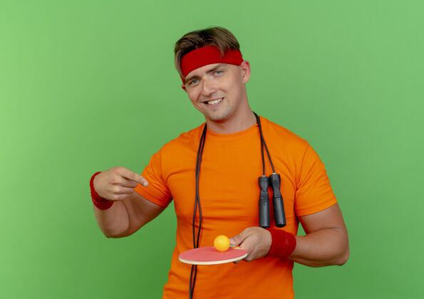 人面带微笑的年轻英俊的运动型男子戴着头带和腕带 脖子上系着跳绳 拿着并指着乒乓球拍 球拍上的球被隔离在绿色的墙上穿着腕带表情