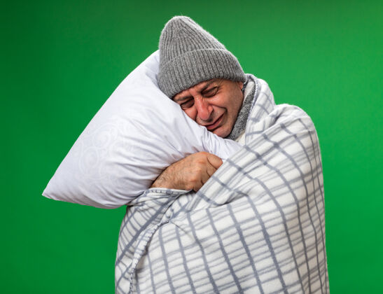 哭哭泣的成年病高加索男子脖子上围着围巾 戴着格子布包裹的冬季帽子 抱着头枕在枕头上 隔离在绿色墙壁上 留着复制空间大人围巾围着