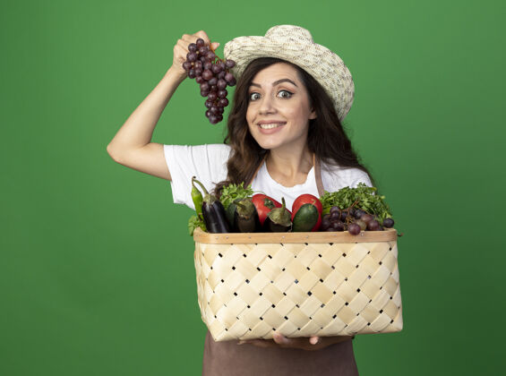 女人年轻的女园丁面带微笑 穿着制服 戴着园艺帽 把菜篮子和葡萄隔离在绿色的墙上表情女人人