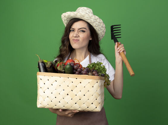人穿着制服的年轻女园丁戴着园艺帽 把菜篮和耙子隔离在绿色的墙上姿势女人篮子