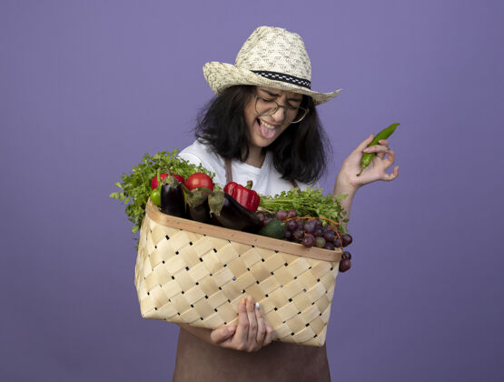 黑发一个年轻的深色皮肤的女园丁戴着眼镜 穿着制服 戴着园艺帽 伸出舌头 把菜篮子和辣椒隔离在紫色的墙上胡椒表情穿着