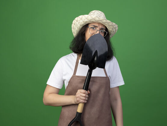 帽子恼怒的年轻黑发女园丁戴着眼镜和制服 戴着园艺帽 站在前面看着隔离在绿色墙上的铁锹黑发表情制服