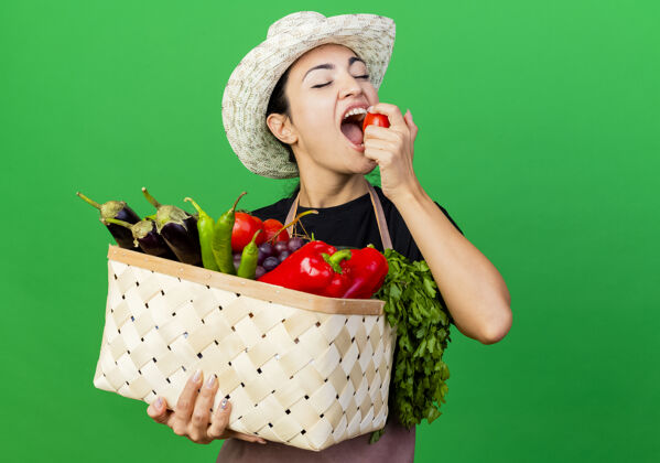 女人年轻漂亮的女园丁围着围裙 戴着帽子 手里拿着装满蔬菜的篮子 咬着西红柿站在绿色的墙上花园脸人