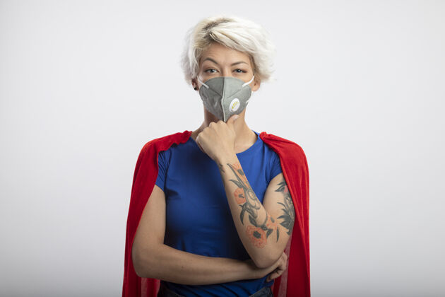 公民自信的女超人身穿红色斗篷 戴着医用面罩 手放在隔离在白墙上的下巴上下巴表情自信